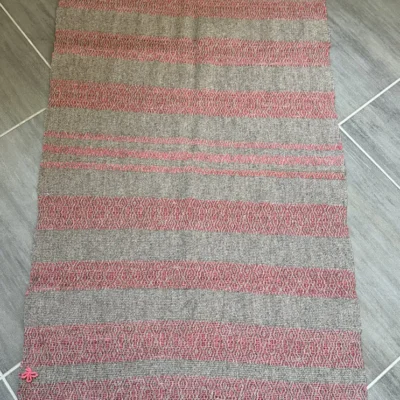 Tapis en laine d'exception Apportez une touche d’authenticité et de chaleur à votre intérieur avec ce tapis d'exception, entièrement tissé à la main sur un métier à bras traditionnel. Une création exclusive signée Line PARERA, fondatrice de L'Échoppe de Linette. Mariage parfait du confort et de l'artisanat d'art, ce tapis valorise la richesse de la filière laine française à travers deux variétés locales : la laine cardée Tarasconnaise au coloris gris naturel et la douceur de la laine cardée Mérinos d'Arles, teinte à l'atelier dans un rouge brique lumineux. Son tissage texturé révèle de subtils motifs géométriques rythmés, sublimés par de jolies finitions frangées. Caractéristiques principales : Matières : 100 % laine française (Tarasconnaise gris naturel et Mérinos d'Arles rouge brique) Teinture artisanale : Faite maison à base de colorants alimentaires certifiés Dimensions : 120 cm x 67 cm (hors franges) Finitions : Franges soignées de 2,5 cm Savoir-faire : Pièce unique exigeant 5 heures de tissage minutieux Idée déco : Idéal en descente de lit, dans une entrée ou pour délimiter un espace cosy avec une pièce de caractère unique.