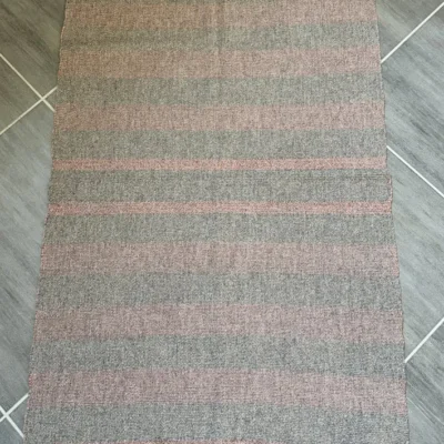 Tapis Artisanal en Laine Locale – Gris Naturel, Marron & Rose Description : Apportez une touche d'authenticité et de chaleur à votre intérieur avec ce tapis unique, entièrement tissé à la main à Toulouse par l'artisane Line Parera. Fruit de 5 heures de travail minutieux sur un métier à bras traditionnel, cette création exclusive associe la texture rustique de la laine cardée Tarasconnaise (en gris naturel) à la douceur exceptionnelle de la laine peignée Mérinos d'Arles. Les bandes marron et rose, délicatement rythmées par un motif géométrique discret, ont été teintées de manière artisanale à l'atelier avec des colorants certifiés. Une pièce unique, durable et écoresponsable, sublimée par de jolies finitions en franges. Caractéristiques : Dimensions : 115 cm x 71 cm (Franges : 8 cm) Composition : 100% laine française (Mérinos d'Arles et Tarasconnaise) Fabrication : Pièce unique tissée main – L'Échoppe de Linette