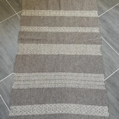 Apportez une touche d'authenticité et de chaleur à votre intérieur avec ce tapis unique, entièrement tissé à la main sur un métier à bras traditionnel par l'artisane Line PARERA. Confectionné à partir de laines locales non teintes, il associe la douceur de la laine cardée mérinos d'Arles (blanc naturel) à la texture authentique de la laine cardée tarasconnaise (grise). Son tissage minutieux révèle un subtil motif géométrique en losanges, sublimé par de généreuses franges torsadées. Caractéristiques : Dimensions : 140 cm × 68 cm (hors franges) Finitions : Franges torsadées de 20 cm Composition : 100% laine cardée locale (Mérinos d'Arles et Tarasconnaise) Fabrication : Artisanale française (8 heures de tissage)