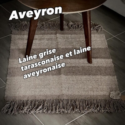 Tapis en Laine Aveyronnaise – Pièce Unique Découvrez l'authenticité de ce tapis artisanal entièrement tissé à la main sur un métier à bras traditionnel par Line PARERA, fondatrice de L'Échoppe de Linette. Une création écoresponsable qui valorise avec passion la filière laine française. Matières d'exception : Laine cardée aveyronnaise (blanc naturel) et laine cardée tarasconnaise (grise). Dimensions : 73 cm x 67 cm. Finitions soignées : Belles franges généreuses de 20 cm et une délicate fleur crochetée en signature. Temps de réalisation : 6 heures de travail minutieux. Apportez une touche chaleureuse, durable et unique à votre intérieur avec ce concentré de savoir-faire local.